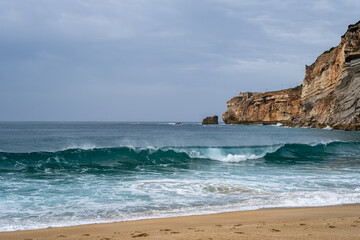 Blue ocean waves rolling onto sandy beach with dramatic coastal cliffs in the background. Natural...