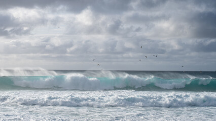 Fototapeta premium Strong wind blows spray off large turquoise waves under a dramatic cloudy sky.