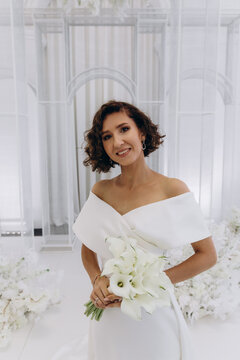 Bride in a flowing white gown holding a flower while posing on an ornate all-white wedding stage with floral decorations. Elegant Bride in White Gown Posing on Luxurious Wedding Stage