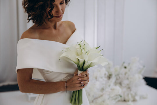 Bride in a flowing white gown holding a flower while posing on an ornate all-white wedding stage with floral decorations. Elegant Bride in White Gown Posing on Luxurious Wedding Stage