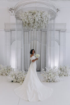 Bride in a flowing white gown holding a flower while posing on an ornate all-white wedding stage with floral decorations. Elegant Bride in White Gown Posing on Luxurious Wedding Stage