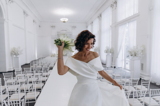 Bride in a flowing white gown holding a flower while posing on an ornate all-white wedding stage with floral decorations. Elegant Bride in White Gown Posing on Luxurious Wedding Stage