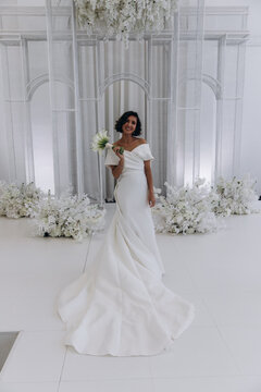 Bride in a flowing white gown holding a flower while posing on an ornate all-white wedding stage with floral decorations. Elegant Bride in White Gown Posing on Luxurious Wedding Stage