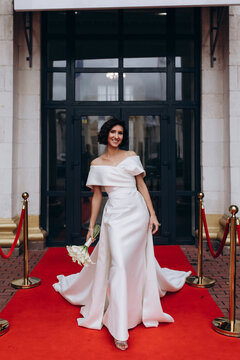 Glamorous bride in an off-shoulder wedding dress holding a calla lily bouquet, posing gracefully on a red carpet in front of a grand building. Elegant Bride on Red Carpet Holding Calla Lily Bouquet