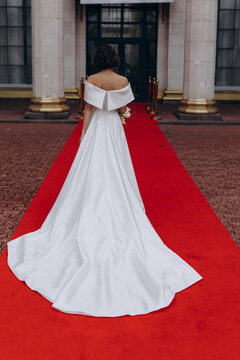 Glamorous bride in an off-shoulder wedding dress holding a calla lily bouquet, posing gracefully on a red carpet in front of a grand building. Elegant Bride on Red Carpet Holding Calla Lily Bouquet