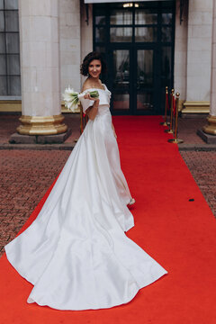 Glamorous bride in an off-shoulder wedding dress holding a calla lily bouquet, posing gracefully on a red carpet in front of a grand building. Elegant Bride on Red Carpet Holding Calla Lily Bouquet
