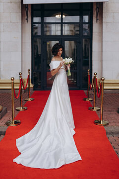 Glamorous bride in an off-shoulder wedding dress holding a calla lily bouquet, posing gracefully on a red carpet in front of a grand building. Elegant Bride on Red Carpet Holding Calla Lily Bouquet