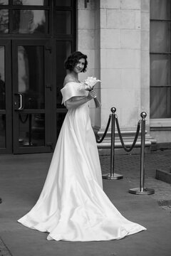Glamorous bride in an off-shoulder wedding dress holding a calla lily bouquet, posing gracefully on a red carpet in front of a grand building. Elegant Bride on Red Carpet Holding Calla Lily Bouquet