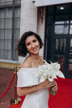 Glamorous bride in an off-shoulder wedding dress holding a calla lily bouquet, posing gracefully on a red carpet in front of a grand building. Elegant Bride on Red Carpet Holding Calla Lily Bouquet