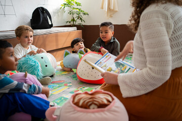 Preschool or kindergarten children sit on the floor listening as the teacher reads a storybook...
