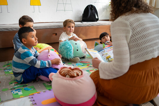 Preschool or kindergarten children sit on the floor listening as the teacher reads a storybook aloud. Concept of early literacy, attention, and storytelling in childhood education