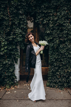 Full-length portrait of a bride in a white gown and leather jacket holding flowers while standing near a lush ivy-covered wall. Bride in Leather Jacket Holding Flowers Near Ivy Wall