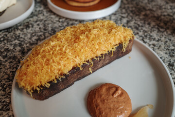 A close-up of a gourmet savory pastry loaf served on a white plate. The dark-crusted bread is generously topped with a thick layer of finely shredded yellow egg and cheese.