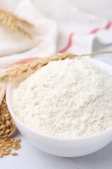 Wheat flour in bowl, grains and spikes on white table, closeup