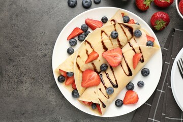 Tasty crepes with strawberries, blueberries and chocolate sauce on gray textured table, flat lay. Space for text