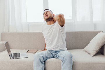 Painful man stretching his neck while sitting on a modern couch. The image captures discomfort,...
