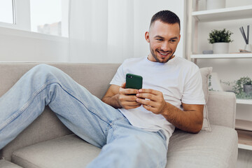 Smiling man relaxing at home, using smartphone with a joyful expression. Bright lighting adds...