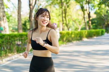 Slim fit Asian woman in sport wear jogs through a park in filled with trees and sunlight.