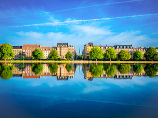 Fototapeta premium Colorful buildings and lush trees reflect on calm waters of Peblinge Sø, Copenhagen, under a vivid blue sky—capturing serene urban beauty in summer.