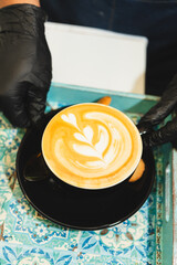 Barista in black gloves serving a cappuccino with latte art in a black cup on a colorful mosaic tray. Artisan coffee moment in a cozy cafe