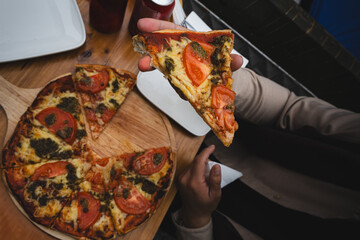 Overhead shot of a person holding a slice of tomato and pesto pizza from a wooden board. Casual dining scene with friends at a cozy café or pizzeria