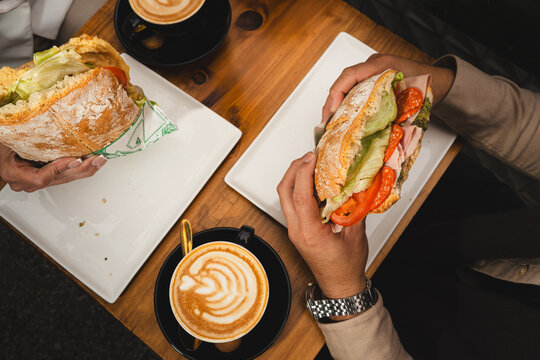 Overhead shot of hands holding fresh sandwiches with cappuccinos on a wooden table. Cozy café brunch with latte art and artisan bread