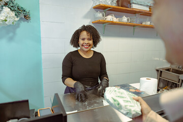 Smiling woman at counter receives wrapped package from customer. Friendly service and bakery interaction in modern kitchen