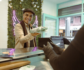 Smiling man receives wrapped food at counter from gloved server in trendy cafe with neon wings...