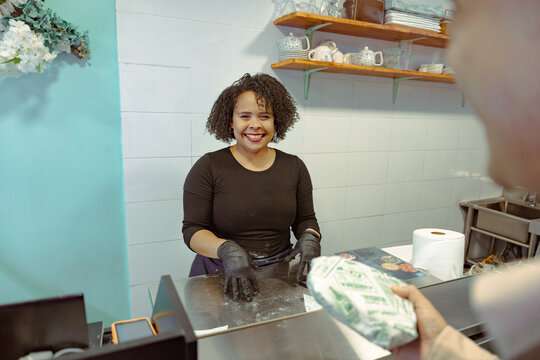 Smiling woman at counter receives wrapped package from customer. Friendly service and bakery interaction in modern kitchen - Powered by Adobe