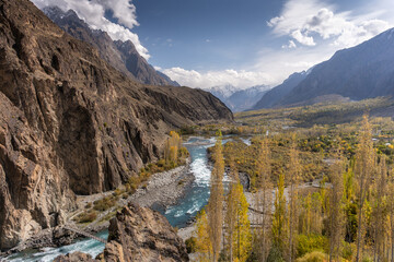 A breathtaking autumn landscape featuring a turquoise river winding through a rocky mountain valley. Golden poplar trees line the banks, and a rustic wooden bridge crosses the water. Snow-capped peaks