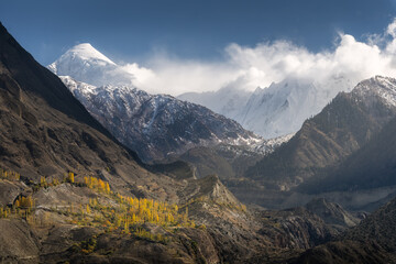 Golden autumn trees adorn the rugged slopes of a dramatic valley. Towering snow-covered peaks rise...