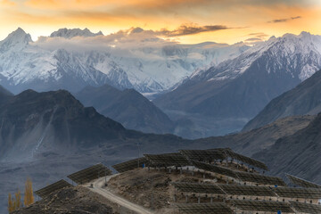 The sun rises behind snowy mountains, painting the sky orange. Below, solar panels line the hillside, blending nature and technology in a peaceful, breathtaking view