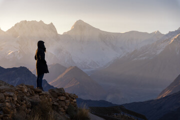 A woman stands on a stone wall at sunrise, gazing at the snow-covered peaks of the mountains. A...