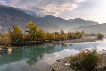 A calm river reflects golden autumn trees and snow-dusted mountains, bathed in soft morning light with mist gently rising in the distance.