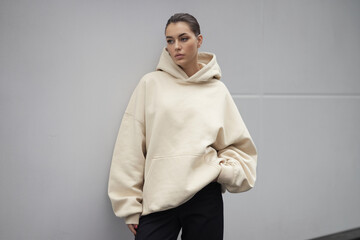 A fashion photography shot of a person wearing an oversized cream-colored hoodie against a plain light gray background. 