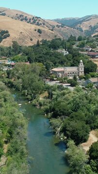 Aerial view of the town of Niles, Fremont, California, nestled in a valley with rolling hills. A river flows through the town, with a dam and a road running alongside it.
