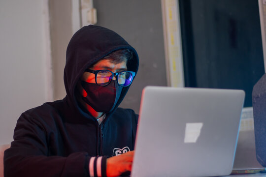 Masked hacker typing on laptop in dark room, surrounded by code screen. Cyber ​​security, identity theft, illegal access, online crime, future digital environment, global threats, innovation and secre