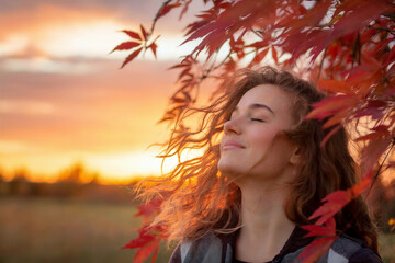 A serene autumn portrait photograph of a young woman with long wavy brown hair against a golden sunset sky. 
