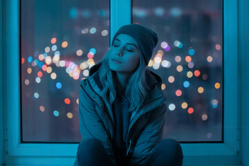 A moody nighttime photograph of a young woman sitting by a window, illuminated by a blue-tinted light. 