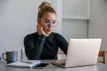 A professional workspace photograph of a young woman wearing a black long-sleeve top and clear-framed glasses, sitting at a white desk.