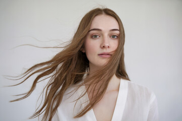 A portrait photograph of a young woman with flowing brown hair against a plain white background. The subject is wearing a white top and has natural, minimal makeup.