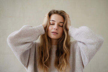 A soft, minimalist portrait photograph of a young woman with long wavy honey-blonde hair against a plain off-white wall. 