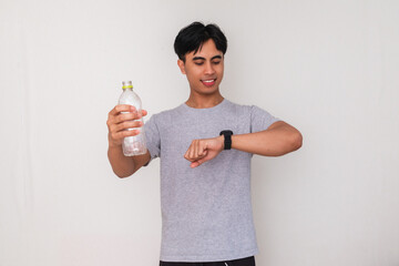 Smiling young Asian man holding a bottle of mineral water while checking his watch, isolated on white background, suitable for advertisement and healthy lifestyle content