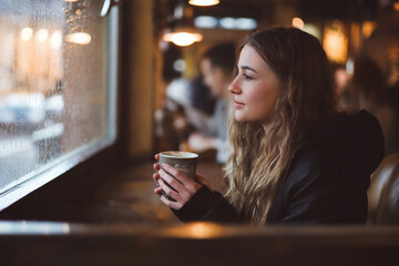 A moody interior photograph of a young woman sitting by a window in a café.
