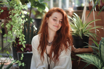 A soft-focused portrait photograph of a young woman with vibrant red wavy hair in a natural indoor setting with plants. She is wearing a flowing white blouse and has a gentle smile.