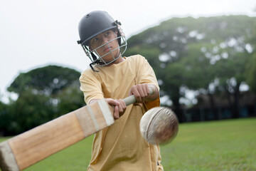 Young boy wearing a helmet plays cricket on a grassy field, focusing intensely as he hits the ball with a wooden bat. Outdoor sports and youth energy captured in motion.