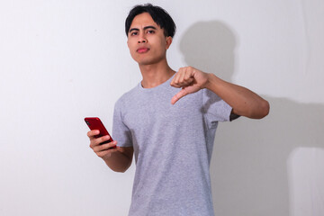 Disappointed young Asian man showing thumbs down while holding a smartphone, isolated on white...