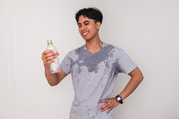 Young Asian man sweating and smiling while holding a water bottle and showing a satisfied gesture,...