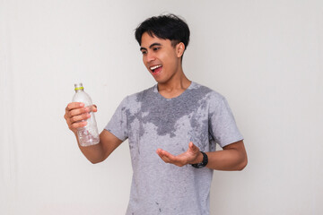 Young Asian man smiling and holding a water bottle with a satisfied expression, sweating after...