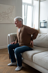 Senior man clutches painful spine unable to stand from couch due to multiple health issues....